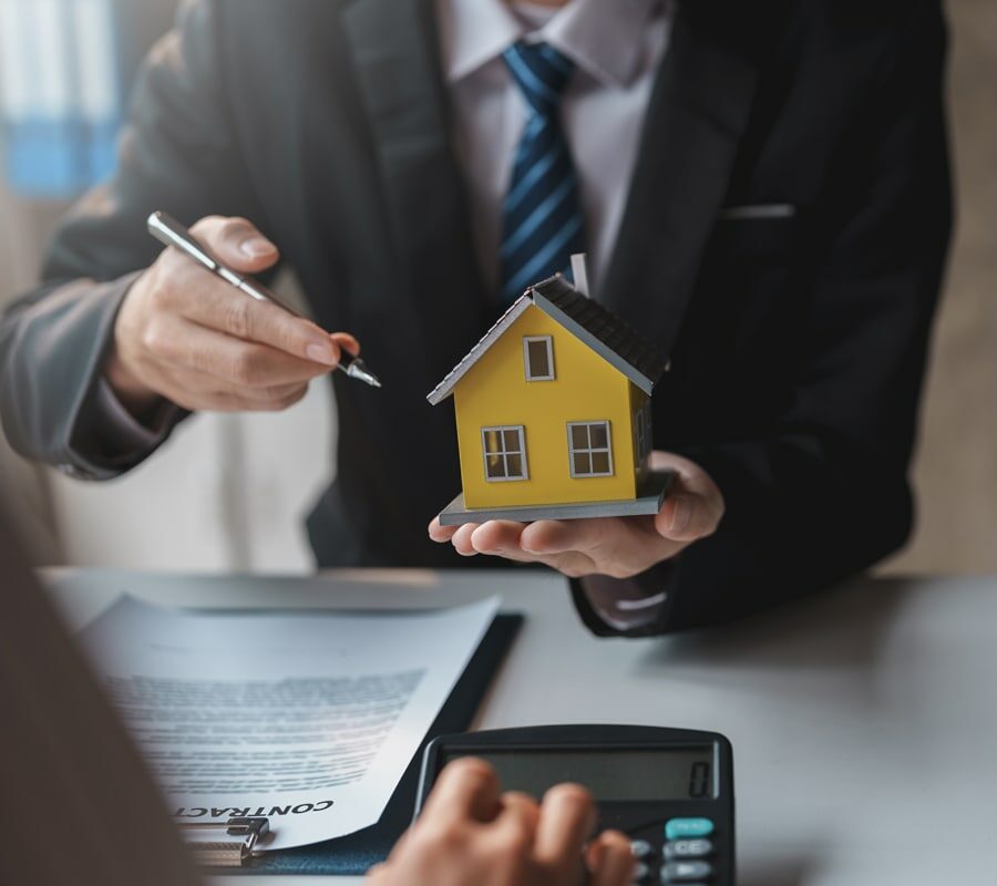 Das Bild zeigt einen Immobilienmakler, der Häuser und Grundstücke an Kunden verkauft, indem er ein Musterhaus als Beispiel zeigt. Es dient als Titelbild der Seite zum Immobilienrecht