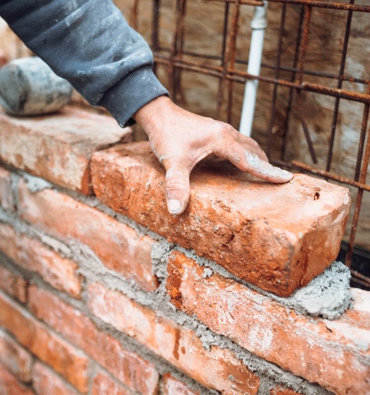 Das Bild zeigt einen Bauarbeiterhand beim Verlegen von Ziegeln für eine Mauer. Es dient als Beitragsbild der Seite zum privaten Baurecht.